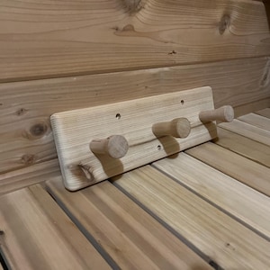 May include: A wooden coat rack with three pegs, mounted on a wooden wall. The rack is made of light-coloured wood. The pegs are cylindrical and extend outwards, providing a space to hang items.