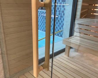 May include: A vertical light-colored wooden door handle with a ribbed texture mounted on a clear glass door. The handle features a rounded top and bottom with a central grip section. The door opens into a sauna with light wood benches and walls.