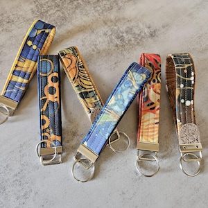 May include: Five colorful fabric keychains with metal key rings. Each keychain has a different pattern and color combination, including blue, yellow, orange, and brown. The keychains are made of fabric and have a quilted texture.