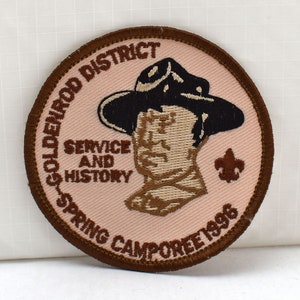 Może przedstawiać: Naszywka haftowana brązowa z beżowym tłem. Naszywka przedstawia kreskówkową postać skauta w kapeluszu z napisem "Goldenrod District Service and History Spring Camporee 1996".