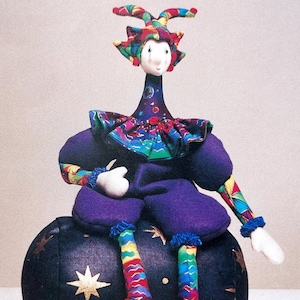 May include: A colorful jester doll with a purple and blue costume sits on a black and gold star patterned ball. The doll has a white face with red cheeks and a long, thin neck.