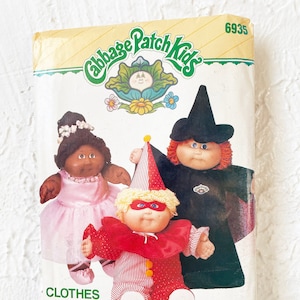 May include: A vintage Cabbage Patch Kids sewing pattern, number 6935, for making clothes for Cabbage Patch Kids dolls. The pattern shows three dolls dressed in a witch costume, a clown costume, and a princess costume. The pattern is titled "Clothes for your Cabbage Patch Kids!" and is by Butterick.