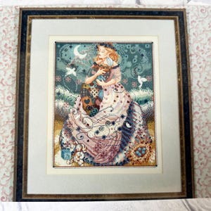 Könnte beinhalten: Gerahmtes Kreuzstich-Kunstwerk mit einer Frau in einem fließenden Kleid, die eine kleine Figur hält. Das Stück hat ein detailliertes, farbenfrohes Design mit einem himmlischen Thema, einschließlich eines Halbmonds und Sternen. Der Rahmen ist dunkel mit goldenen Akzenten.