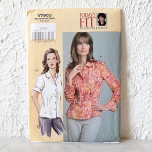 May include: A vintage sewing pattern labelled "V7903" with images of women's blouses. The pattern cover shows a woman in a floral blouse and another in a white blouse. The text "TODAY'S FIT BY SANDRA BETZINA" is also visible.