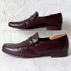 May include: A pair of dark burgundy leather penny loafers featuring a classic moc-toe construction, a horizontal strap across the vamp, and a low stacked heel. The shoes have a smooth, polished finish and include wooden shoe trees inside each.