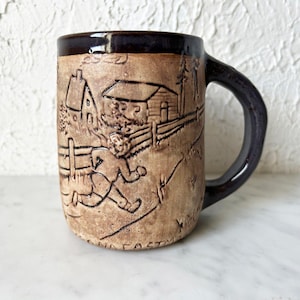 Puede incluir: Una taza de cerámica marrón con borde y asa marrón oscuro. La taza presenta un detallado relieve de una escena rural con una casa, una valla y una figura corriendo. La taza mide aproximadamente 10 cm de alto.