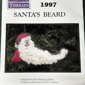 May include: A white felt Santa beard ornament with a red hat and a white face. The ornament is on a green background. The text "THREADS 1997 SANTA'S BEARD" is printed on the card. "Original artwork by Emerson Quillin Needlearts adaptation by Ruth A. Sparrow" is printed below the ornament.
