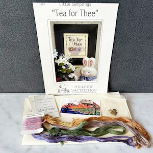 May include: A cross-stitch kit with the text "Tea for Thee" and a framed cross-stitch design of a teacup. The kit includes embroidery floss in various colors, a ceramic bunny figurine, and a small card.