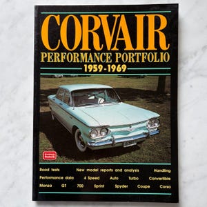 May include: A book titled Corvair Performance Portfolio 1959-1969 with a black cover featuring large yellow text and a photograph of a light blue vintage car parked on grass. The cover includes text listing road tests, model reports, and car models.