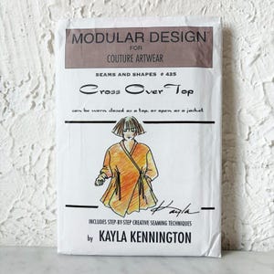 May include: A white sewing pattern envelope titled Modular Design for Couture Artwear Seams and Shapes #425 Cross Over Top by Kayla Kennington. The front features a hand-drawn illustration of a wrap-style top with ties in orange and yellow.
