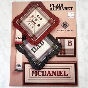 May include: A vintage "Plaid Alphabet" cross-stitch pattern book. The cover features examples of cross-stitched designs, including a pillow with the word "DAD", a framed "B", and a framed "McDaniel" nameplate. The designs incorporate plaid patterns and decorative borders.