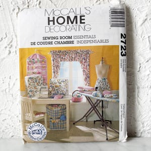 May include: A McCall's Home Decorating sewing pattern envelope. The image shows a sewing room scene with various essentials, including a sewing machine, ironing board, and fabric storage. The text on the envelope reads "Sewing Room Essentials".
