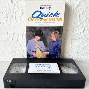 Puede incluir: Una cinta VHS negra con una etiqueta blanca que dice "sewing with nancy" y "Quick Gifts & Decor". La etiqueta también incluye el texto "60 MIN. 37846" y "P.O. Box 683, Beaver Dam, WI 53916". La cinta está etiquetada como "T-65".