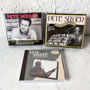 Op de afbeelding: Drie cd-hoesjes met Pete Seeger. Op het bovenste linkerhoesje staat "PETE SEEGER" en "WE SHALL OVERCOME". Op het bovenste rechterhoesje staat "PETE SEEGER SING-A-LONG". Op het onderste hoesje staat "PETE SEEGER LIVE AT NEWPORT".