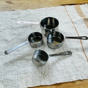 May include: Four silver measuring cups of different sizes on a white and brown striped kitchen towel.