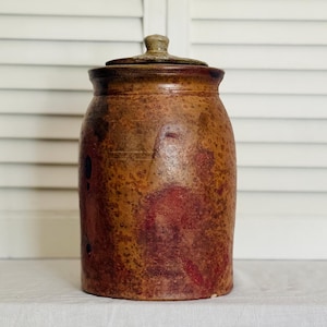 May include: A vintage brown stoneware jar with a lid. The jar has a rounded body and a slightly textured surface with reddish-brown and tan hues. The lid has a small knob. The jar is likely used for storage.