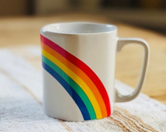 May include: White ceramic mug with a rainbow design. The rainbow is made up of red, orange, yellow, green, blue, indigo, and violet stripes.