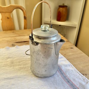May include: Vintage aluminum camping kettle with a copper handle and glass knob on the lid. The kettle is cylindrical with a spout and sits on a white cloth with red and blue stripes. A classic piece for outdoor enthusiasts.