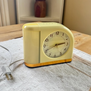 May include: A vintage, pale yellow electric alarm clock with a cream-colored clock face. The clock has a curved design with a wood-tone base and a ribbed side panel. The clock face displays the time with black numerals and hands.
