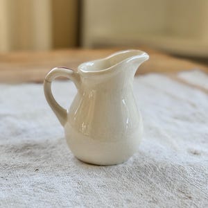 May include: A small, white ceramic creamer with a handle. The creamer has a textured surface and a slightly rounded shape.