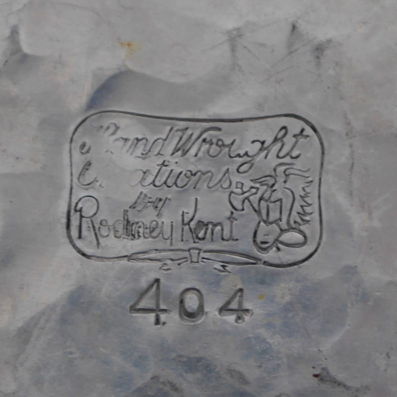 Peut inclure: Gros plan d'une &eacute;tiquette en m&eacute;tal argent&eacute; avec le texte "Hand Wrought Creations by Rodney Kent" et un motif d'oiseau stylis&eacute;. Le num&eacute;ro "404" est &eacute;galement visible sous l'&eacute;tiquette.