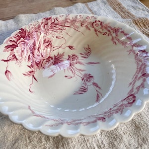 May include: A white ceramic bowl with a scalloped edge and a pink floral design. The bowl is decorated with a large floral motif in the center and a smaller floral border around the rim.