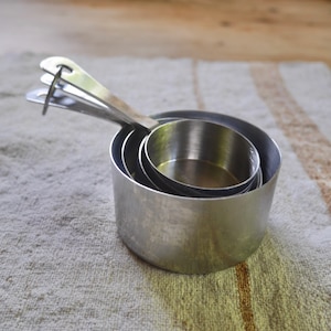 May include: A set of four nested silver measuring cups. The cups are made of stainless steel and have a shiny finish.