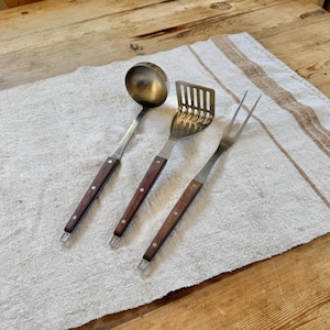 May include: A set of vintage kitchen utensils, including a ladle, slotted spatula, and fork, with dark wood handles and stainless steel heads. The tools are arranged on a light-colored linen cloth with brown stripes, set on a wooden table.