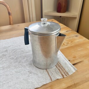 May include: A vintage aluminum coffee pot with a clear glass knob on the lid and a black handle. The coffee pot is sitting on a beige and brown striped cloth on a wooden table. A wooden chair and shelf are in the background.
