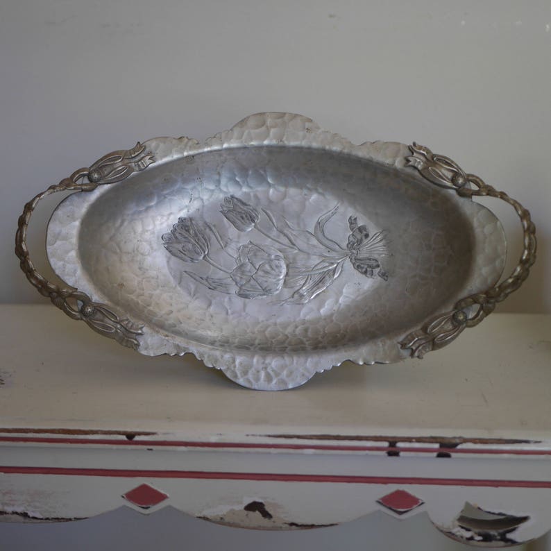 Peut inclure: Un plateau de service ovale en argent avec deux poign&eacute;es. Le plateau a une finition martel&eacute;e et un motif floral en relief sur le fond.