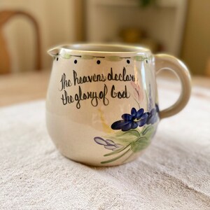 May include: A beige ceramic pitcher with a handle, featuring a floral design with blue, purple, and yellow flowers. The pitcher has the text "The heavens declare the glory of God" written in black cursive.