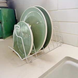May include: A white wire dish rack holds three green plates with cream-colored rims. The plates are arranged on a light-colored countertop near a white tiled wall and a white sink.