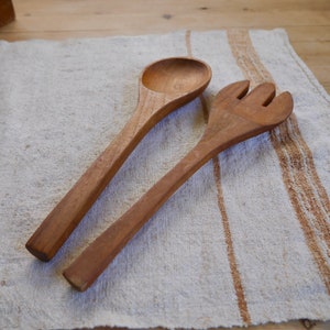 May include: A wooden salad serving set with a spoon and a fork. The set is on a white and brown striped linen cloth.