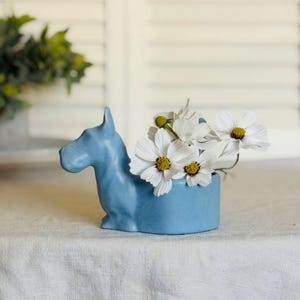 May include: A light blue ceramic dog figurine with a white flower arrangement inside. The dog is sitting on a white tablecloth.