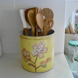 May include: A yellow metal utensil holder with a floral design featuring crackers. The holder is filled with wooden spoons, spatulas, and a fork.