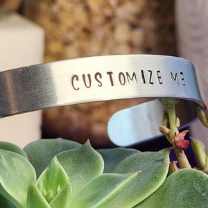 May include: A silver-colored cuff bracelet with the words "CUSTOMIZE ME" stamped on the front. The bracelet is resting on a green succulent plant. The background includes jars and a white pot.