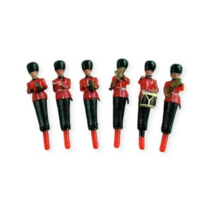 May include: Set of six miniature toy soldiers dressed in red uniforms with black hats and holding various musical instruments. The soldiers are standing on a white background.