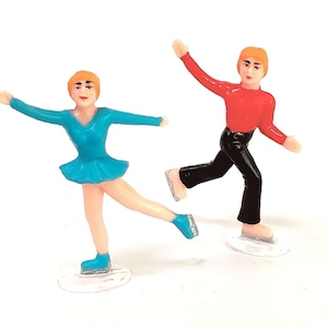 May include: Two small plastic ice skater figurines. One wears a teal dress, the other a red top and black trousers. Both are on clear plastic bases and have orange hair.
