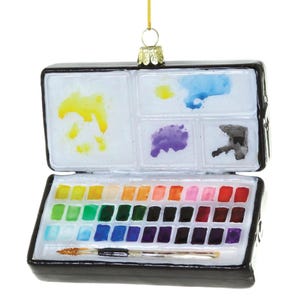 May include: A black rectangular watercolor paint set ornament featuring an open lid with mixing wells containing yellow, blue, purple, and black paint. The base holds rows of various colored paint pans and a small paintbrush with a gold tip.