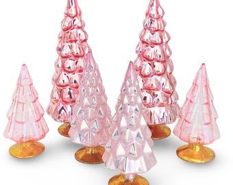 May include: A set of six pink glass Christmas trees with gold bases. The trees are all different sizes and shapes, and they are arranged in a cluster on a white background.