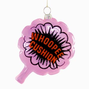 May include: A pink, novelty Christmas ornament shaped like a whoopee cushion. The ornament has a black outline with the words "WHOOPEE CUSHION" in orange letters. A gold string is attached for hanging.