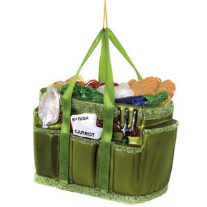 May include: A green glass ornament shaped like a gardening tote bag. The tote is filled with colorful vegetables and gardening tools. A tag reads "Radish Carrot". The ornament has green handles and glitter accents.