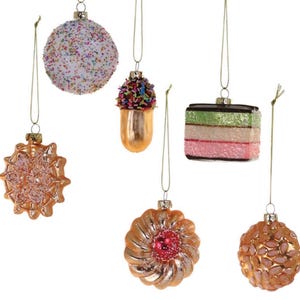May include: A collection of hanging Christmas ornaments in various shapes and colors. The ornaments include a round one covered in sprinkles, a gold acorn shape, a layered cake, and several flower-shaped ornaments. Each ornament has a gold string for hanging.