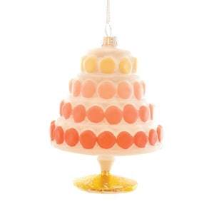 May include: A tiered cake ornament with a white base and layers of orange, pink, and yellow circular decorations. The ornament is suspended by a silver string and has a gold-colored base. This festive decoration is perfect for the holidays.