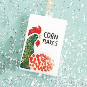 Op de afbeelding: Een witte rechthoekige kerstbal met een illustratie van een haan en de woorden "CORN FLAKES" in het zwart. De haan is groen en goud met een rode kam en lel. De bal hangt aan een gouden draad.