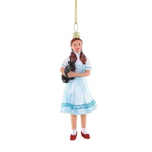 May include: A decorative ornament depicting a character from The Wizard of Oz. The figure wears a light blue dress, white blouse, and red shoes, holding a black dog. The ornament is suspended by a gold-colored string.