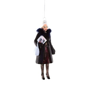 May include: A glass ornament of a woman wearing a black coat with a blue fur collar, black tights, and black heels. She is holding a magazine that says "RUNWAY" and a white shopping bag.