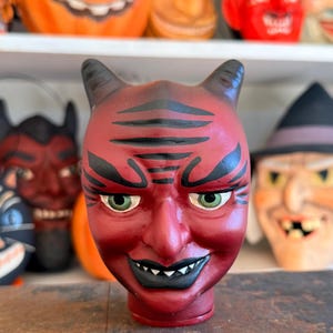May include: A red devil head figurine featuring black painted eyebrows and forehead lines, green eyes, and a black mouth with white teeth. The figure has two small horns on top of the head and a smooth, rounded base, presented as a standalone object.