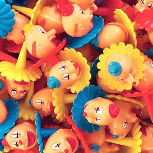 May include: A bowl filled with colorful clown-themed cake toppers. The toppers feature red, yellow, blue, and white clown faces with different expressions. Each topper has a small plastic stick for inserting into a cake.