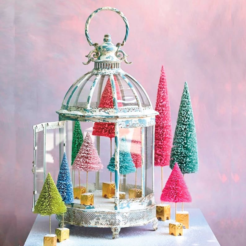 May include: A distressed white metal lantern with a hinged door containing several miniature bottle brush trees in shades of pink, blue, green, and red. Each tree stands on a small gold square base, creating a colorful display of festive decor.